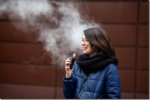Vape teenager. Young pretty white caucasian girl in a blue jacket and glasses smoking an electronic cigarette opposite modern brown background on the street in the winter. Deadly bad habit.